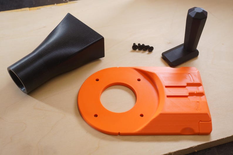 May include: An assortment of black and orange 3D-printed parts, likely for a woodworking tool. The components include a black nozzle, an orange base with a circular opening, a black handle, and small black screws. The parts are arranged on a light-colored wooden surface.