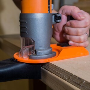 May include: An orange and gray power router being used on a piece of wood. The router has a clear plastic dust collection port and is connected to a black hose. The tool is being held by a person.