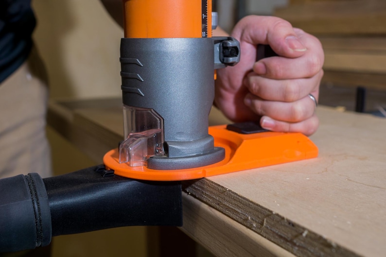 May include: An orange and gray power router being used on a piece of wood. The router has a clear plastic dust collection port and is connected to a black hose. The tool is being held by a person.