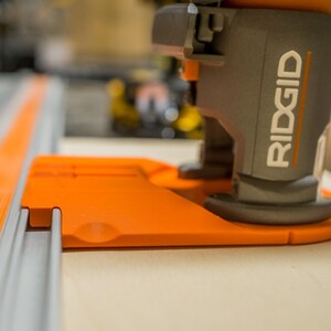 May include: Close-up of a gray and orange woodworking tool with the brand name "RIDGID" visible. The tool is positioned on a light-colored wooden surface, with a gray track and orange accents in the foreground.