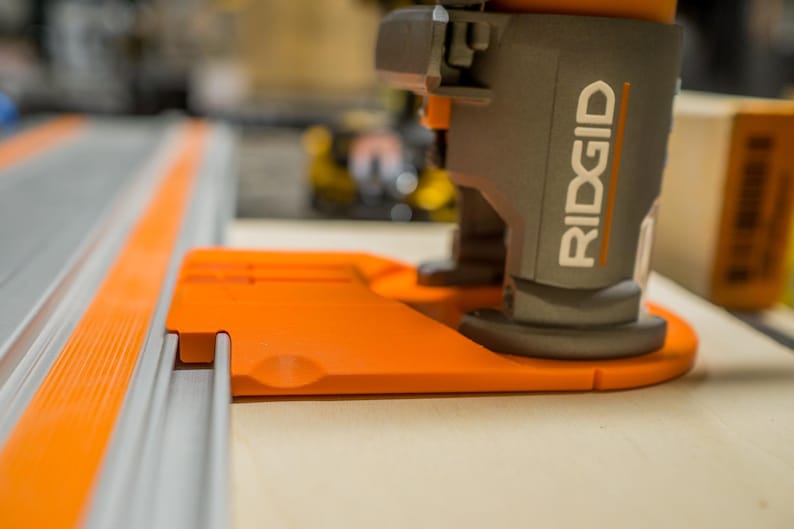 May include: Close-up of a gray and orange woodworking tool with the brand name "RIDGID" visible. The tool is positioned on a light-colored wooden surface, with a gray track and orange accents in the foreground.