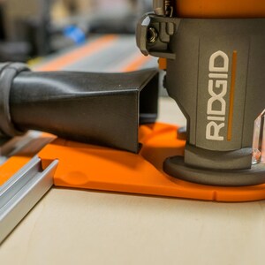 May include: Close-up of a gray and orange RIDGID power tool. The tool has a black hose attached and is mounted on an orange base. The word "RIDGID" is printed in white on the side of the tool.
