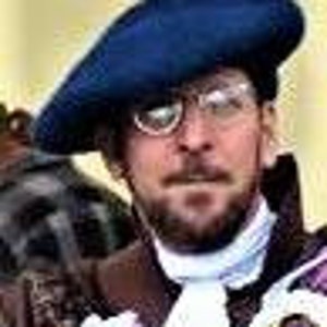 May include: A man wearing a dark blue beret, round eyeglasses, and a patterned jacket with a white ruffled collar. He has a beard and mustache. The background is blurred, suggesting an outdoor setting.