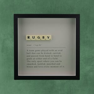 Peut inclure: Un cadre carré noir de type boîte avec un fond blanc présentant le mot RUGBY formé par des tuiles de lettres crème. Sous les tuiles se trouve une définition imprimée de style dictionnaire du sport, le décrivant comme un jeu d'équipe.