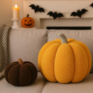 Può includere: Due cuscini a forma di zucca in peluche, arancioni e marroni, su un divano grigio. La zucca più grande è arancione brillante con un gambo verde, mentre quella più piccola è marrone scuro con un gambo marrone chiaro. Decorazioni di Halloween.