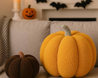 May include: Two plush pumpkin pillows in shades of orange and brown sit on a gray couch. The larger pumpkin is a bright orange with a green stem, while the smaller one is dark brown with a tan stem. Halloween decor.
