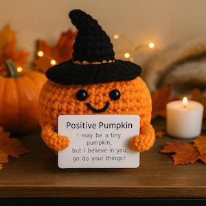 Peut inclure: Une citrouille orange crochetée portant un chapeau de sorcière noir, tenant une pancarte avec l'inscription "Positive Pumpkin". La pancarte dit "I may be a tiny pumpkin, but I believe in you, go do your things !" Une bougie allumée et des feuilles d'automne sont en arrière-plan.