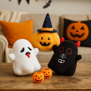 Könnte beinhalten: Eine Sammlung von Halloween-Plüschtieren, darunter ein weißer Geist, ein Kürbis mit Hexenhut und ein schwarzer Teufel. Zwei kleine orangefarbene Kürbis-Kerzenhalter sind davor. Die Spielzeuge stehen auf einem Holztisch.