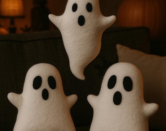 May include: Three white felt ghost decorations with black oval eyes and mouths. One ghost is floating above the other two. The background includes a pumpkin, a bat silhouette, and a lamp.