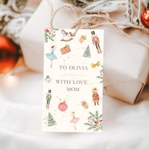 Puede incluir: Una etiqueta de regalo rectangular con un diseño de acuarela que presenta una bailarina, un cascanueces y un árbol de Navidad. La etiqueta dice "TO OLIVIA WITH LOVE, MOM". La etiqueta está atada a un regalo envuelto con hilo.