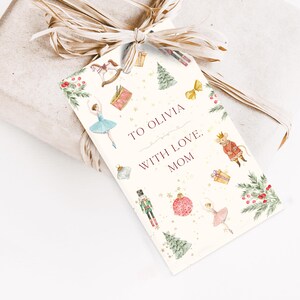 May include: A gift tag with a watercolor design featuring a ballerina, nutcracker, and Christmas tree. The tag reads "TO OLIVIA WITH LOVE, MOM" and is tied to a wrapped gift with a raffia bow.