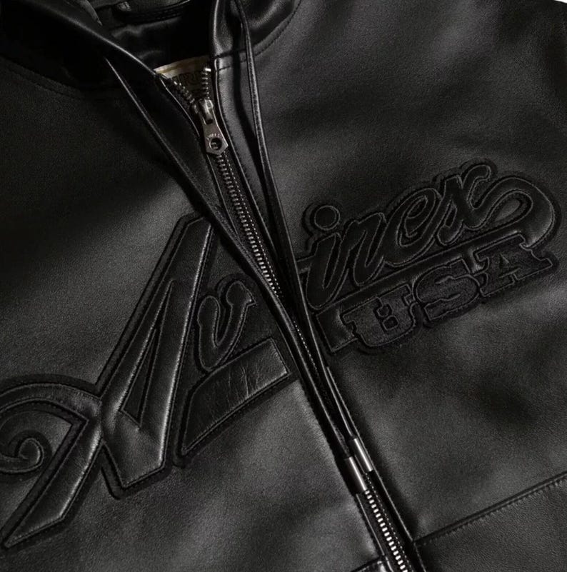 May include: Black leather jacket with a zip closure. The jacket features the embroidered word "A&shy;virex USA" in a stylized font on the front. The jacket has a classic design.