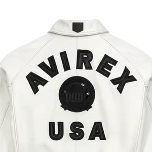 May include: White leather jacket with black embroidered "AVIREX" lettering and a circular emblem. Below the emblem, the word "USA" is embroidered in black. The jacket has a classic collar and a small emblem on the back of the collar.