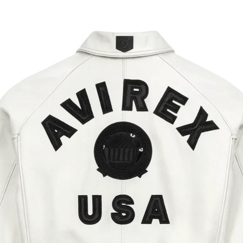 May include: White leather jacket with black embroidered "AVIREX" lettering and a circular emblem. Below the emblem, the word "USA" is embroidered in black. The jacket has a classic collar and a small emblem on the back of the collar.