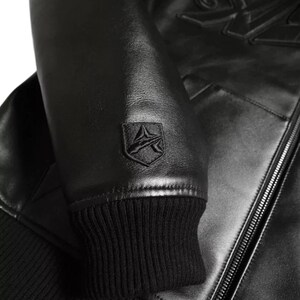May include: Black leather jacket featuring a ribbed knit cuff and a small emblem on the sleeve. The jacket has a zip closure and a smooth, shiny finish. The emblem is a stylized design within a shield shape.