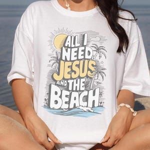 May include: White t-shirt with the text "ALL I NEED JESUS AND THE BEACH" in a graphic design featuring a sun, palm trees, and waves. The shirt is worn by a person wearing sunglasses and a blue bandana.