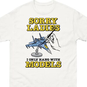 May include: White t-shirt with a graphic design featuring a blue model airplane being painted by a hand. The text reads "SORRY LADIES I ONLY HANG WITH MODELS" in yellow block letters. A humorous design for model enthusiasts.