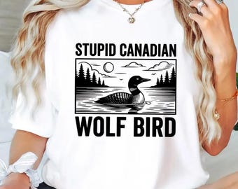 May include: White t-shirt with a black and white graphic of a loon on water, with trees, a moon, and the text "STUPID CANADIAN WOLF BIRD". The shirt has a crew neck and short sleeves.
