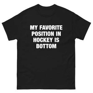 May include: Black t-shirt with white text that reads "MY FAVORITE POSITION IN HOCKEY IS BOTTOM." The shirt has short sleeves and a crew neck. The text is centered on the front of the shirt.