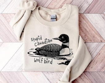 May include: Beige sweatshirt with a black and white graphic of a loon bird on water, with the text "Stupid Canadian Wolf Bird". The sweatshirt is folded with the sleeves rolled up.
