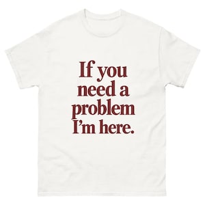 May include: A white short-sleeved t-shirt featuring the dark red text "If you need a problem I'm here." printed in a centered, serif font on the chest. The garment has a crew neckline and a standard fit, displaying clear, bold lettering.