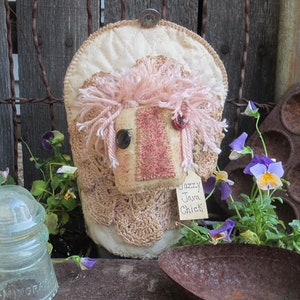 May include: A handmade, rustic, cream-colored, crocheted chick ornament with pink yarn hair and a red button nose. The ornament is hanging on a wooden fence with a tag that reads "Jazzy Java Chick".