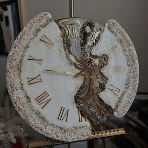 Peut inclure: Une horloge ronde décorative avec un cadran blanc et des chiffres romains dorés. Le centre de l'horloge présente un motif marron et doré, et le bord extérieur est orné de cristaux scintillants. L'horloge est exposée sur un chevalet doré.