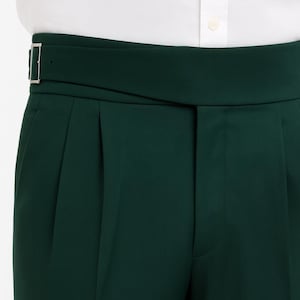 May include: Close-up of dark green trousers with a wide waistband and a silver buckle. The trousers have pleats and a tailored fit. A white shirt is visible at the top of the frame.