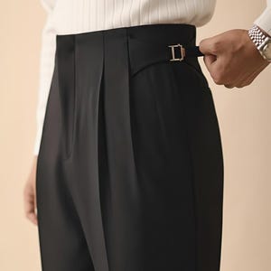 Op de afbeelding: Zwarte broek met hoge taille en een goudkleurige gesp. De broek heeft een geplooide voorkant en een getailleerde pasvorm. De persoon draagt een wit geribbelde longsleeve.