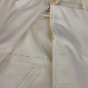 Puede incluir: Tela de color crema, posiblemente seda o sat&eacute;n, con un brillo sutil. La tela est&aacute; doblada y drapeada, mostrando su textura y un borde crudo sin terminar. Se ven finas costuras azules.