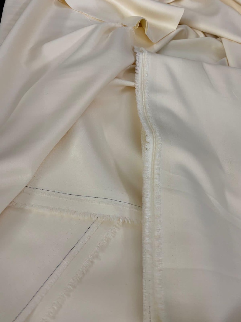 Puede incluir: Tela de color crema, posiblemente seda o sat&eacute;n, con un brillo sutil. La tela est&aacute; doblada y drapeada, mostrando su textura y un borde crudo sin terminar. Se ven finas costuras azules.