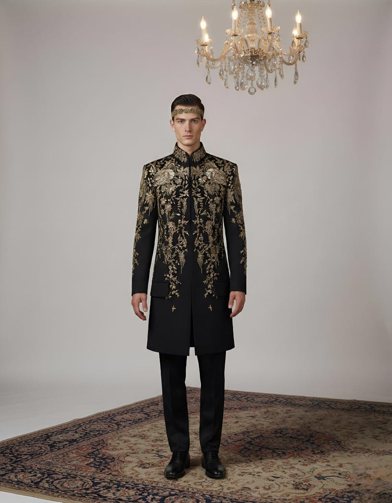 May include: A black coat with gold floral embroidery, paired with black pants and shoes. The coat features intricate gold detailing on the front and sleeves. A gold headband is worn. A chandelier hangs in the background. The outfit is formal.