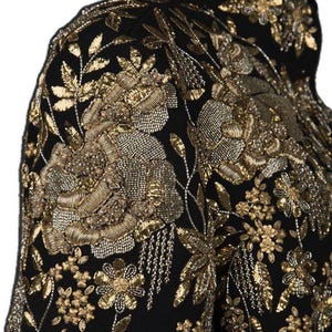 May include: Close-up of a black jacket with intricate gold embroidery. The jacket features floral and leaf patterns, with detailed beadwork and metallic thread creating a luxurious texture. The design covers the shoulder and upper chest area.