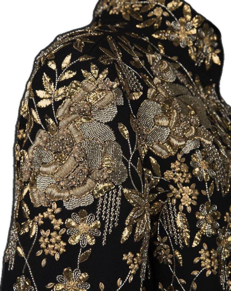 May include: Close-up of a black jacket with intricate gold embroidery. The jacket features floral and leaf patterns, with detailed beadwork and metallic thread creating a luxurious texture. The design covers the shoulder and upper chest area.