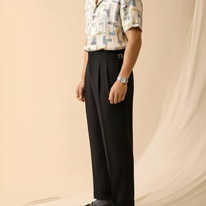 Puede incluir: Una camisa de manga corta con un estampado geom&eacute;trico en beige, azul y amarillo se combina con pantalones negros plisados. El conjunto se completa con mocasines de cuero marr&oacute;n. Se ve un reloj plateado en la mu&ntilde;eca.