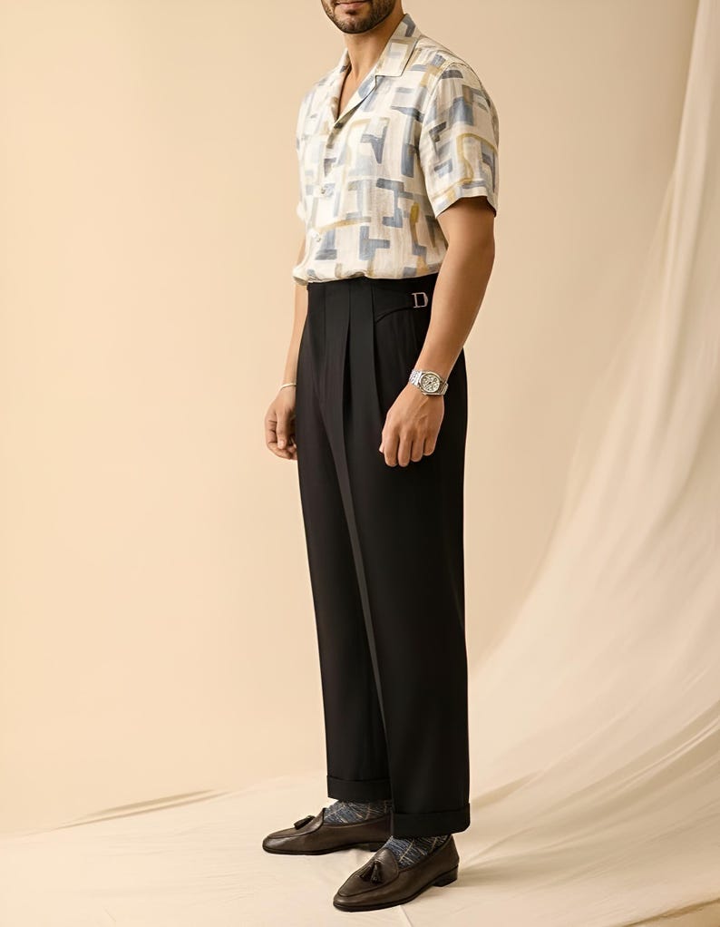 Puede incluir: Una camisa de manga corta con un estampado geom&eacute;trico en beige, azul y amarillo se combina con pantalones negros plisados. El conjunto se completa con mocasines de cuero marr&oacute;n. Se ve un reloj plateado en la mu&ntilde;eca.