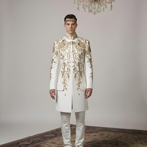 May include: A formal white suit with intricate gold embroidery. The long jacket features a mandarin collar and is paired with matching trousers. The outfit is accessorized with a gold headband. The background includes a chandelier and a patterned rug.