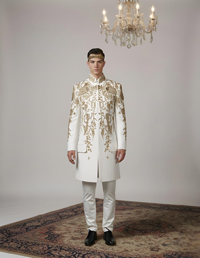 May include: A formal white suit with intricate gold embroidery. The long jacket features a mandarin collar and is paired with matching trousers. The outfit is accessorized with a gold headband. The background includes a chandelier and a patterned rug.