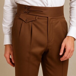 May include: Close-up of brown high-waisted trousers with a wide waistband and pleats. The trousers have a buttoned pocket and three decorative buttons on the waistband. The trousers are paired with a white dress shirt.