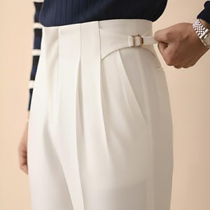 Puede incluir: Primer plano de un pantal&oacute;n blanco plisado con cintura alta y detalle de hebilla. El pantal&oacute;n se combina con un jersey de punto acanalado azul marino. Una mano ajusta la hebilla del pantal&oacute;n.