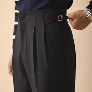Puede incluir: Primer plano de un pantal&oacute;n negro con hebilla dorada. El pantal&oacute;n tiene la parte delantera plisada y un corte a medida. La persona lleva una camisa de rayas azul marino y blanco.