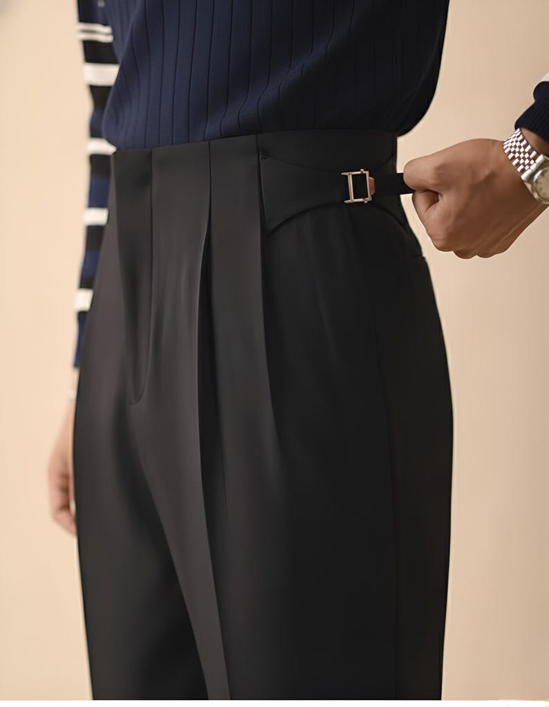 Puede incluir: Primer plano de un pantal&oacute;n negro con hebilla dorada. El pantal&oacute;n tiene la parte delantera plisada y un corte a medida. La persona lleva una camisa de rayas azul marino y blanco.