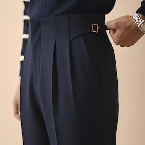 Puede incluir: Pantalones plisados azul marino con cintura alta y hebilla dorada. Los pantalones tienen un corte a medida y un dise&ntilde;o cl&aacute;sico. La persona lleva una parte superior azul marino con una manga a rayas.