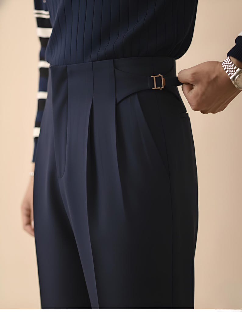 Puede incluir: Pantalones plisados azul marino con cintura alta y hebilla dorada. Los pantalones tienen un corte a medida y un dise&ntilde;o cl&aacute;sico. La persona lleva una parte superior azul marino con una manga a rayas.