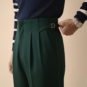 Puede incluir: Pantalones plisados ​​verde oscuro con hebilla dorada. Los pantalones tienen cintura alta y corte entallado. La imagen muestra un primer plano de la cintura, destacando el dise&ntilde;o y la tela.