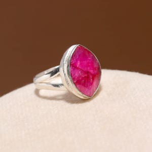 Peut inclure: Une bague en argent ornée d'une grande pierre précieuse rose vif de forme marquise. La pierre est sertie dans un sertissage à lunette, et la bague a un design à tige fendue. La bague est présentée sur une surface crème.