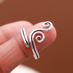 Puede incluir: Un anillo plateado con un diseño único y arremolinado. El anillo presenta un diseño abierto que se envuelve alrededor del dedo, con dos adornos en espiral. El anillo se muestra en un dedo sobre un fondo borroso.