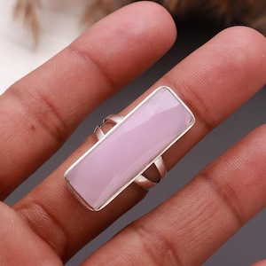 May include: A rectangular ring featuring a smooth, light pink gemstone set in a polished silver-toned metal band. The stone has a flat, elongated surface that spans across the finger, held securely by a simple, minimalist metal frame setting.