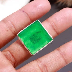 Peut inclure: Une bague de forme carrée dotée d'une pierre précieuse verte translucide et vibrante, sertie dans une bande en métal argenté poli. La pierre présente une surface facettée avec des inclusions internes visibles et une teinte vert émeraude.