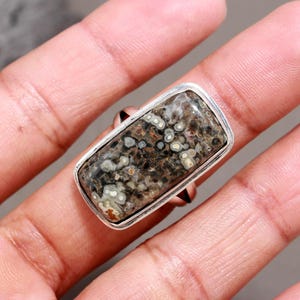 May include: A rectangular ring featuring a polished stone with a complex pattern of dark brown, black, and cream-coloured circular inclusions. The stone is set within a smooth, silver-toned metal bezel that frames the rectangular shape of the gemstone.